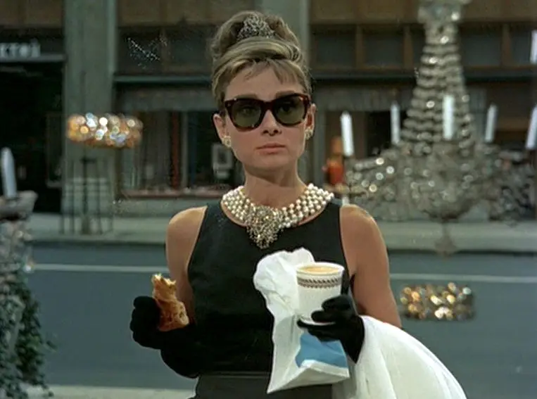 Stylish woman in a black dress, pearl necklace, and sunglasses on a city street, holding a coffee and a pastry with gloves on.